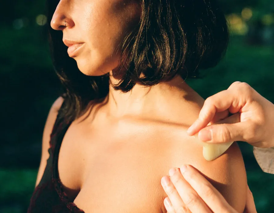 Une praticienne passe une plaque de gua sha sur l’épaule d’une personne, en extérieur devant un feuillage
