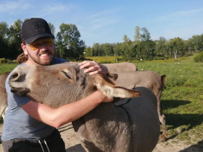 Josh Whelan, casquette et lunettes, un bras autour d’un âne dans un pré