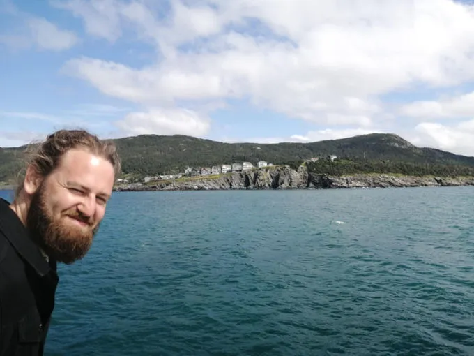 Josh Whelan souriant au bord de l’eau, un village côtier et des falaises en arrière-plan