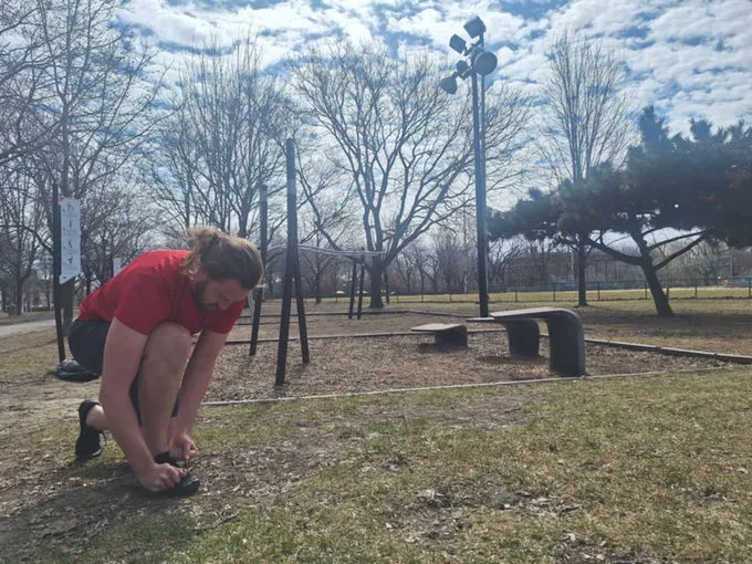 Josh Whelan en tenue de sport, laçant ses chaussures dans un parc urbain au printemps